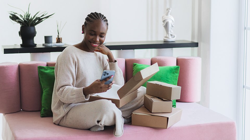 Woman sitting on a couch with packages stacked next to her which she smiles at her phone.